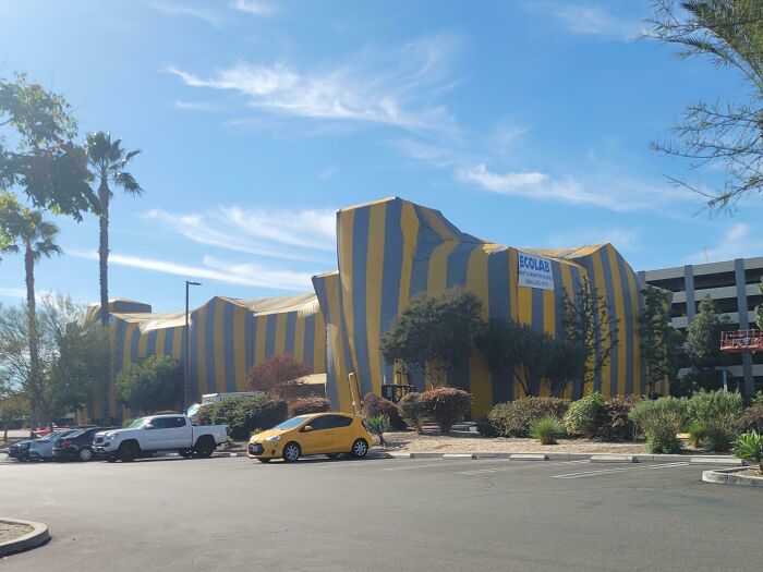 Edificio envuelto en telas amarillas y negras en un estacionamiento con autos y árboles bajo un cielo azul claro.