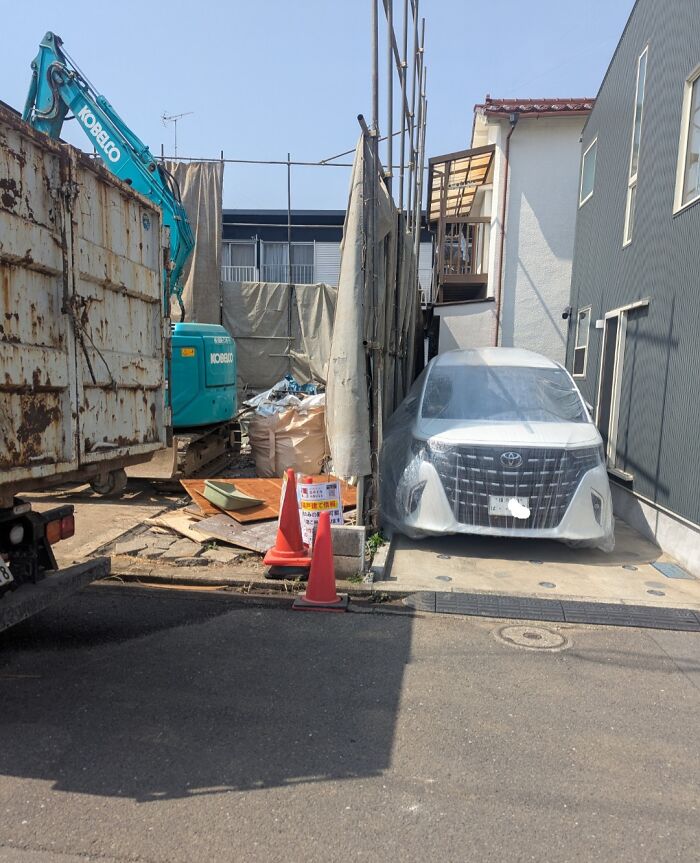 Coche cubierto con plástico en obra de construcción entre edificios, mostrando soluciones a problemas habituales en espacios pequeños.