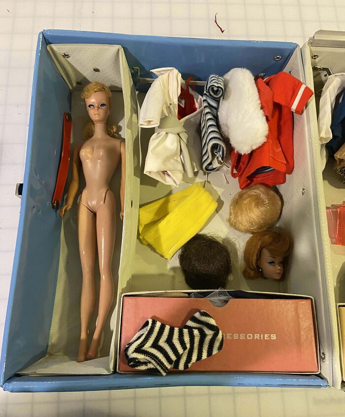 Muñeca vintage sin ropa junto a accesorios y pelucas organizados en caja mostrando perspectiva temporal.