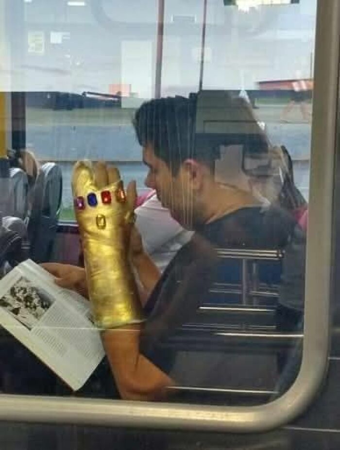 Hombre en transporte público leyendo un libro con un guante dorado gigante y coloridas gemas, imagen curiosa de personas.