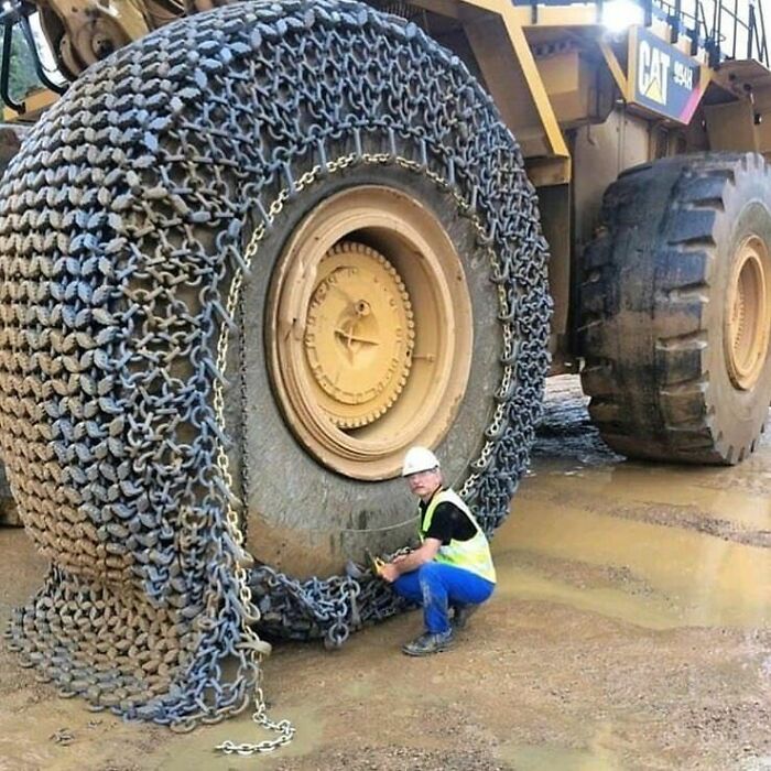 Hombre con casco ajustando cadenas en rueda gigante de maquinaria pesada en terreno embarrado, foto interesante y curiosa.