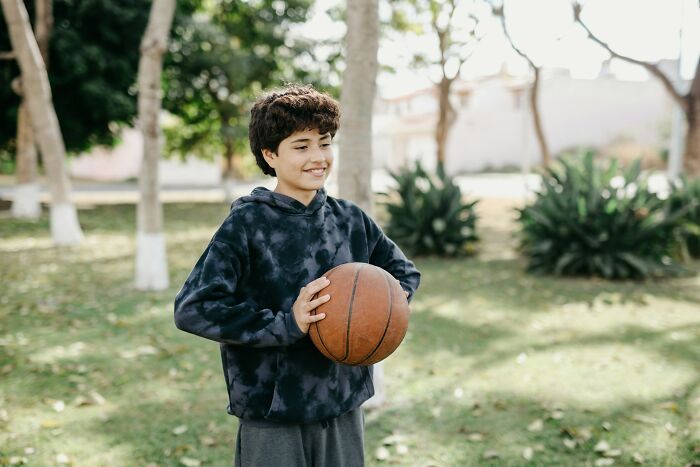 Niño con genes buenos sonriente sosteniendo una pelota de baloncesto en un parque durante el día soleado.