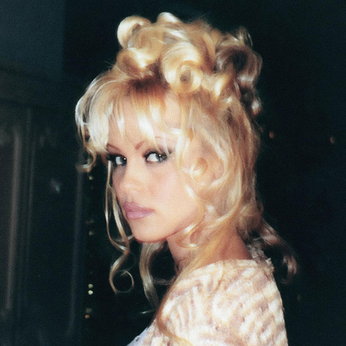 Pamela Anderson con estilo glamuroso de los 90, maquillaje retro y cabello rubio voluminoso con rizos definidos.