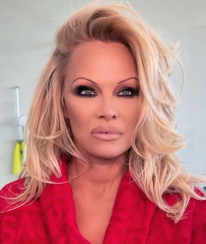 Pamela Anderson con estilo glamuroso de los 90, maquillaje de ojos audaz y cabello rubio voluminoso en bata roja.
