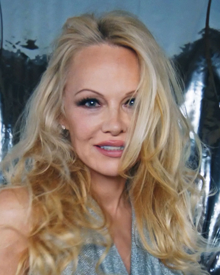 Pamela Anderson con look glamuroso de los 90, maquillaje llamativo y cabello rubio voluminoso en ambiente elegante.