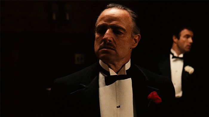 Hombre con esmoquin oscuro y rosa roja en solapa, retrato en ambiente oscuro de celebridad problemática.