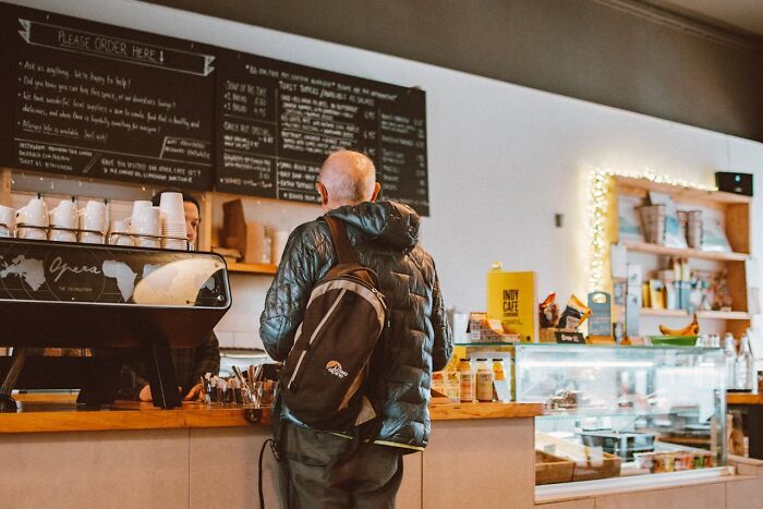 Hombre con mochila en cafetería pidiendo café, ilustrando trucos vitales muy útiles en su día a día cotidiano.