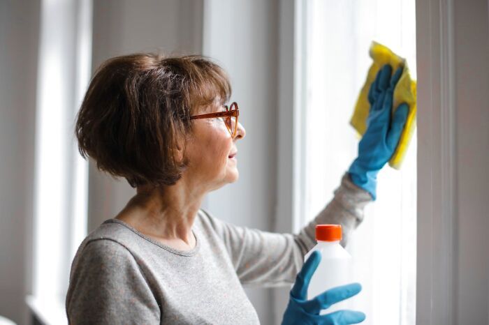 Mujer usando trucos vitales para limpiar una ventana, con guantes azules y un paño amarillo en casa.