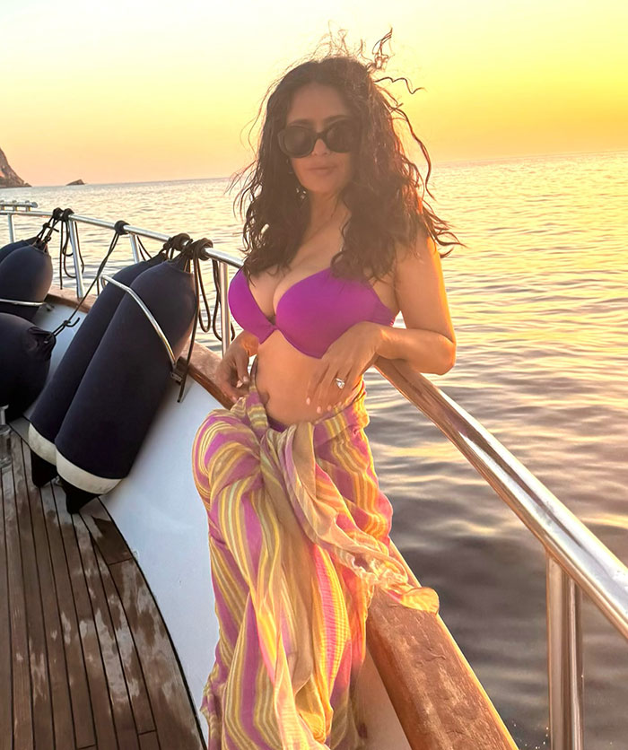 Salma Hayek en bikini morado y pareo a rayas posando en un bote al atardecer para Sports Illustrated Swimsuit.