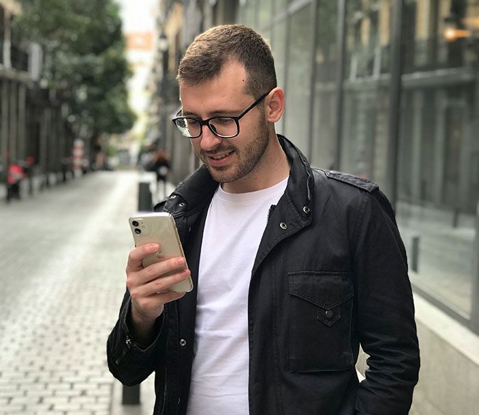 Hombre con gafas y chaqueta negra revisando su teléfono en la calle, posible señal de infidelidad pasada por alto.