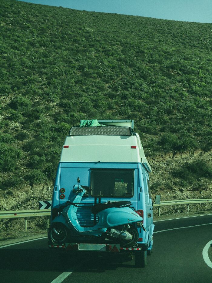 Furgoneta azul con scooter en carretera rodeada de paisaje verde, mostrando combinación única y lotería genética.