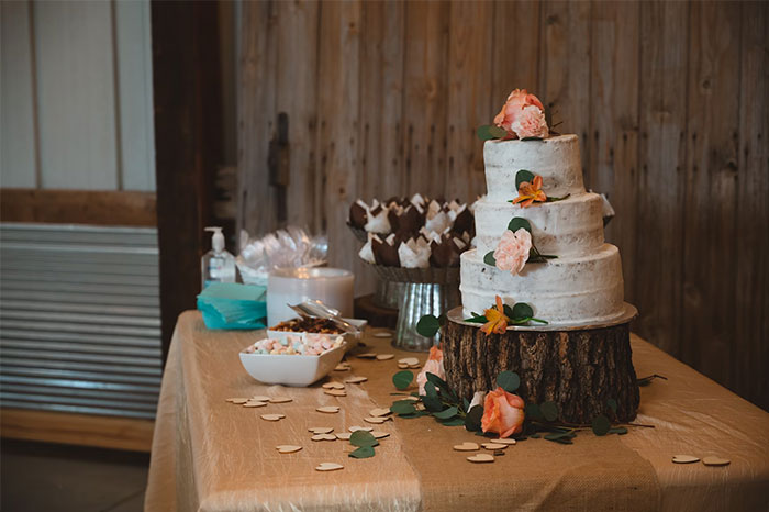 Pastel de bodas rústico con flores decorativas sobre base de madera en mesa con postres y decoración sencilla.