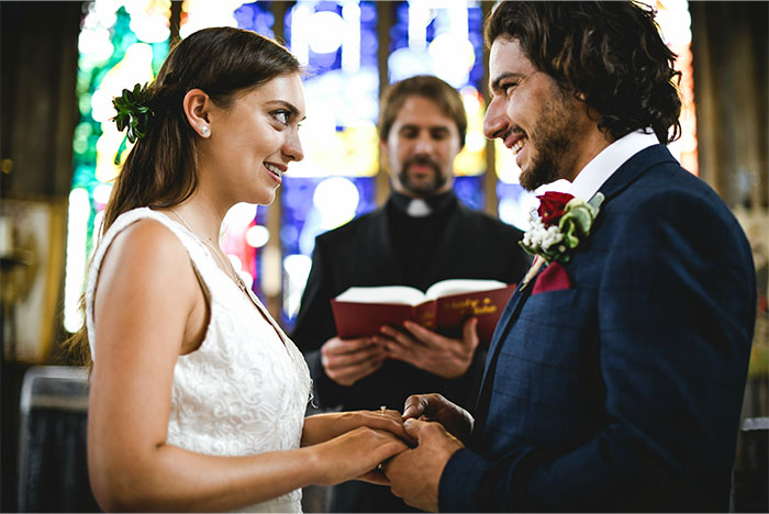 Pareja de novios intercambiando anillos durante ceremonia de boda en iglesia, con sacerdote leyendo texto sagrado.
