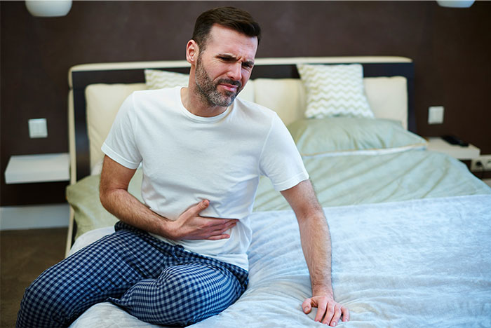 Hombre con pijama sentado en cama sosteniéndose el estómago, ilustrando historias de bodas que fueron de mal en peor.