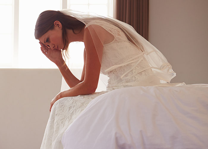 Novia triste sentada en cama con vestido y velo blanco en una habitación iluminada, reflejando una boda que salió mal.