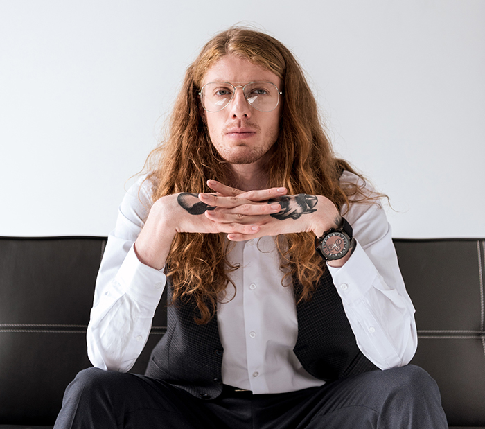 Hombre joven con cabello largo y tatuajes en las manos, mostrando rasgos de genética fuerte y única sentado en sofá.