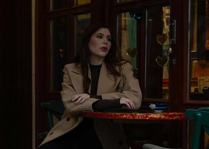 Mujer pensativa en cita terribles, sentada en mesa de restaurante con abrigo beige y fondo con decoración de corazones.