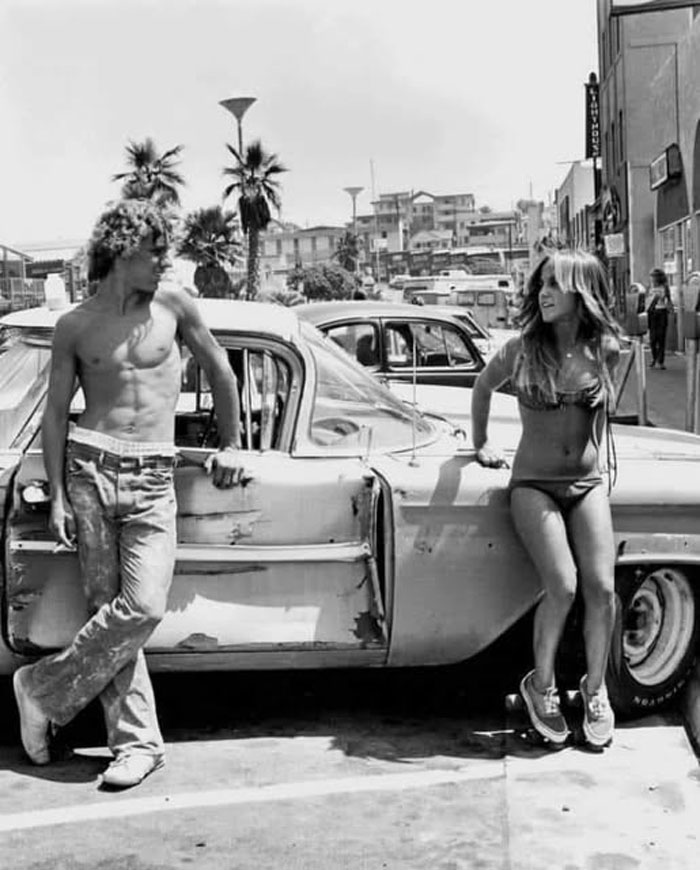 Pareja joven junto a un coche antiguo, foto histórica curiosa mostrando moda y vida urbana del pasado inédito.