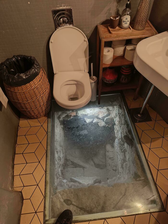 Baño con inodoro y suelo de cristal que muestra restos arqueológicos, imagen aterradora e inquietante en un ambiente cerrado.
