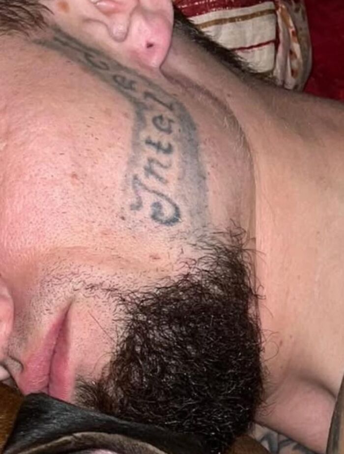 Tatuaje facial mal hecho con letras borrosas en la mejilla de un hombre con barba oscura y piel clara.