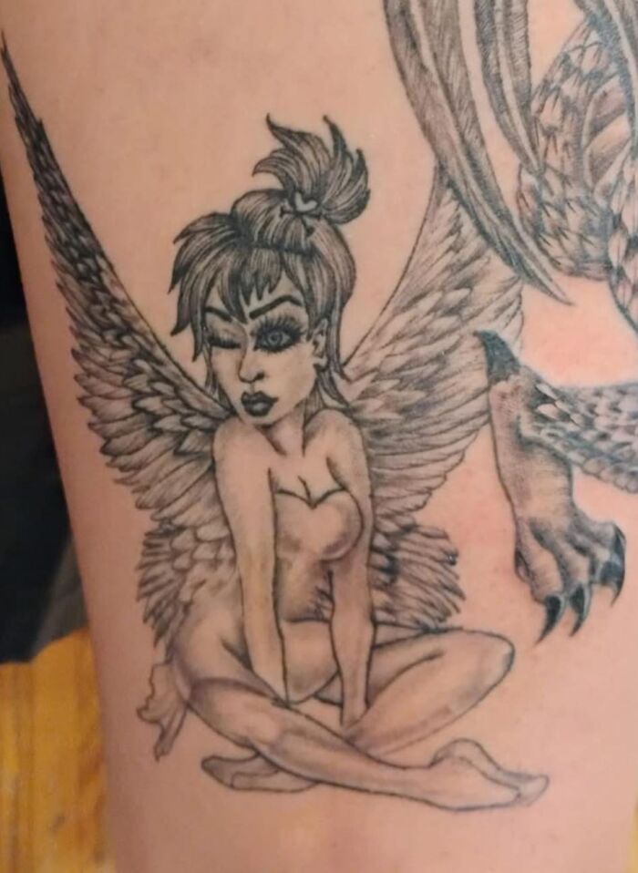 Tatuaje feo de hada con alas grandes y pose sentada en pierna con expresión guiñando un ojo, ejemplo de tatuajes horribles.