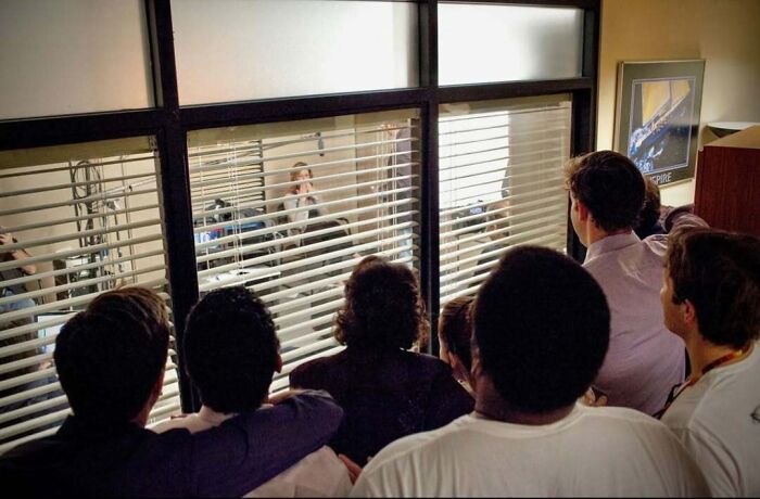 Grupo de personas observando a través de una ventana con persianas, mostrando ángulos alternativos en un evento interior.