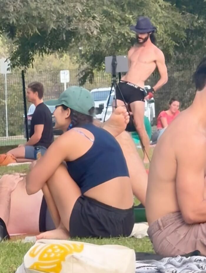 Hombre influencer de pie con sombrero y short negro causando sensación en parque donde varias personas están sentadas.