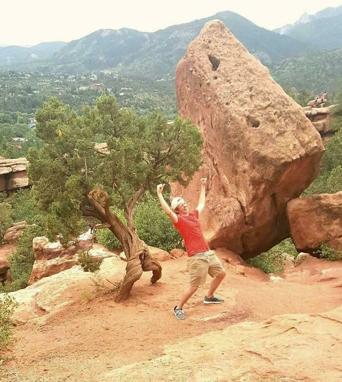 Hombre posando divertido con roca grande en equilibrio, foto divertida para hacerte reír en paisaje montañoso.