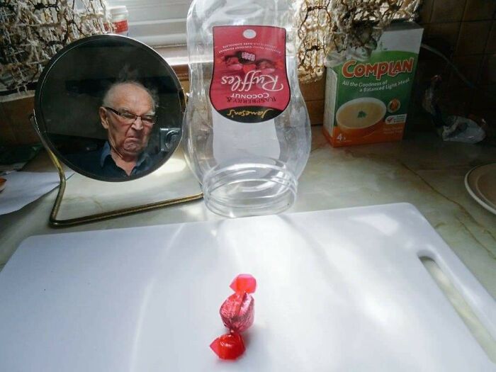 Hombre mayor con expresión divertida reflejada en espejo junto a dulce rojo sobre superficie blanca en cocina.