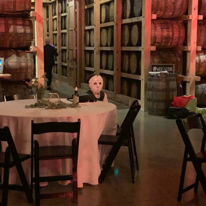 Niño con máscara de terror sentado en una mesa de fiesta en bodega con barriles, imagen divertida para hacerte reír hoy.