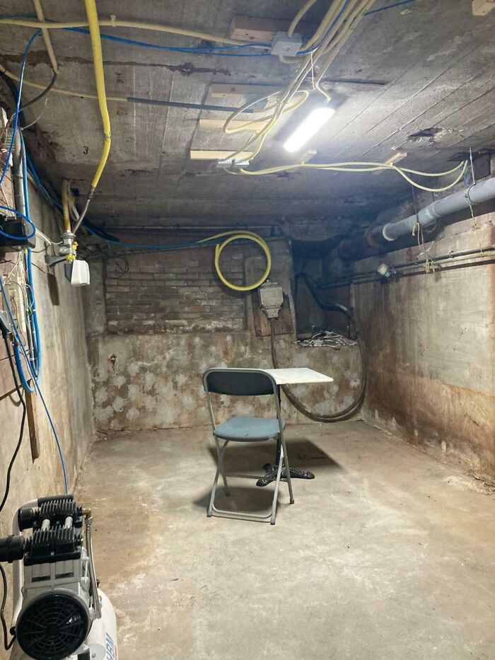 Lugar de trabajo tóxico en sótano con paredes de concreto, cables expuestos y mobiliario improvisado en espacio descuidado.