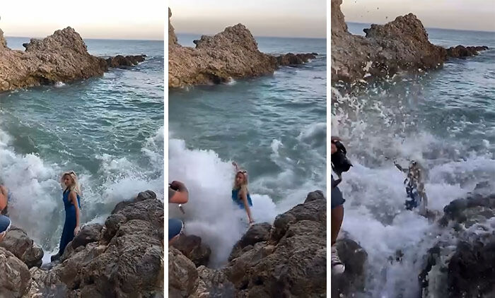 Influencia vergonzosa de mujer en vestido azul sorprendida por ola mientras posa en rocas junto al mar.