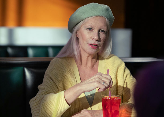 Mujer mayor en restaurante con boina verde y suéter amarillo disfrutando una bebida roja, escena de citas terribles presenciadas.