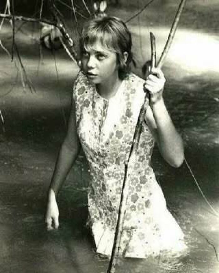 Niña en vestido floreado cruzando un río, sosteniendo una rama, foto histórica mostrando curiosidades del pasado.