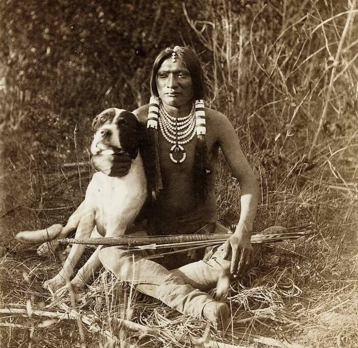 Hombre indígena con perro y armas, foto histórica que muestra curiosidades culturales y tradiciones ancestrales.