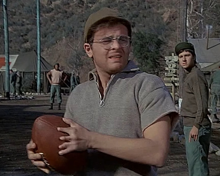 Hombre con gorra y gafas sostiene un balón en campo militar, destacando peculiaridades corporales de celebridades.