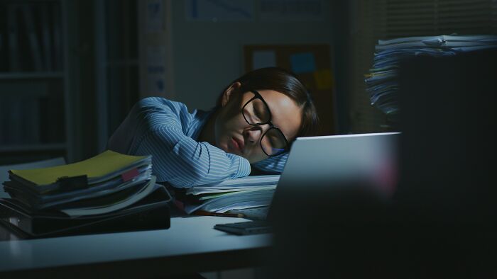 Mujer terapeuta agotada durmiendo sobre escritorio con pilas de documentos y laptop en ambiente oscuro trabajo terapias.