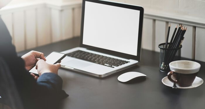 Persona usando teléfono frente a una laptop con pantalla en blanco, en escritorio con taza de café y lápices, experiencias online.