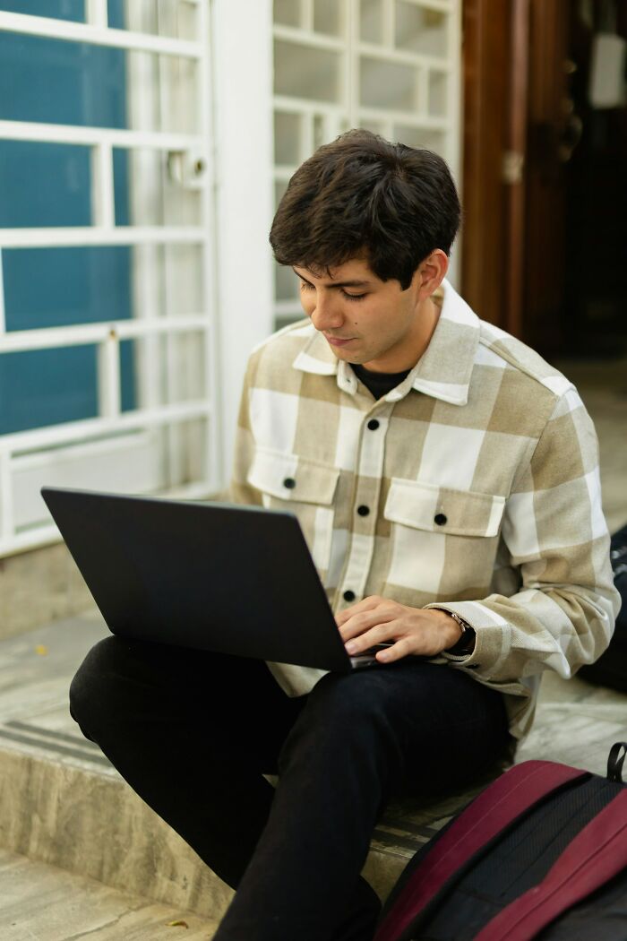 Joven sentado en escaleras con laptop, ilustrando momentos de corregir a la persona equivocada y fallar miserablemente.