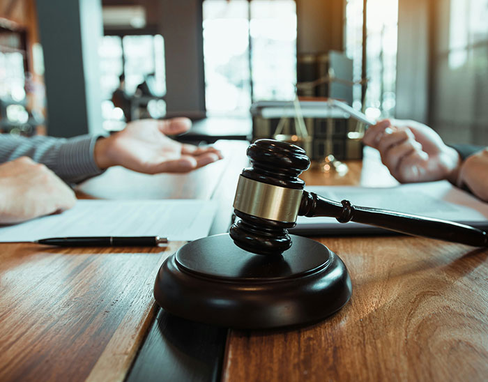 Mazo de abogado en primer plano mientras dos personas discuten documentos legales en una mesa de madera, casos de abogados.