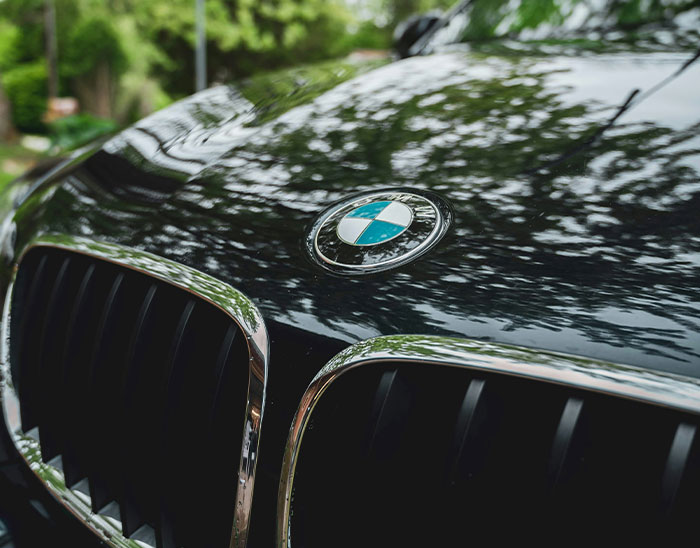 Detalle del logo BMW en capó negro de auto con reflejos de árboles, relacionado con abogados y clientes.