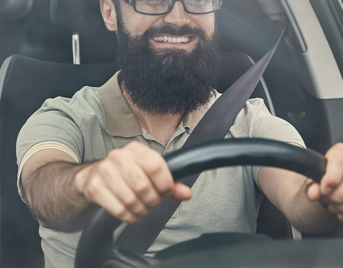 Hombre con barba sonriendo mientras conduce un coche usando cinturón de seguridad, relacionado con abogados y clientes.