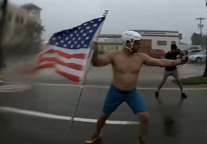Hombre con casco y bandera estadounidense en clima lluvioso, ilustrando diferencias en longevidad entre mujeres y hombres.