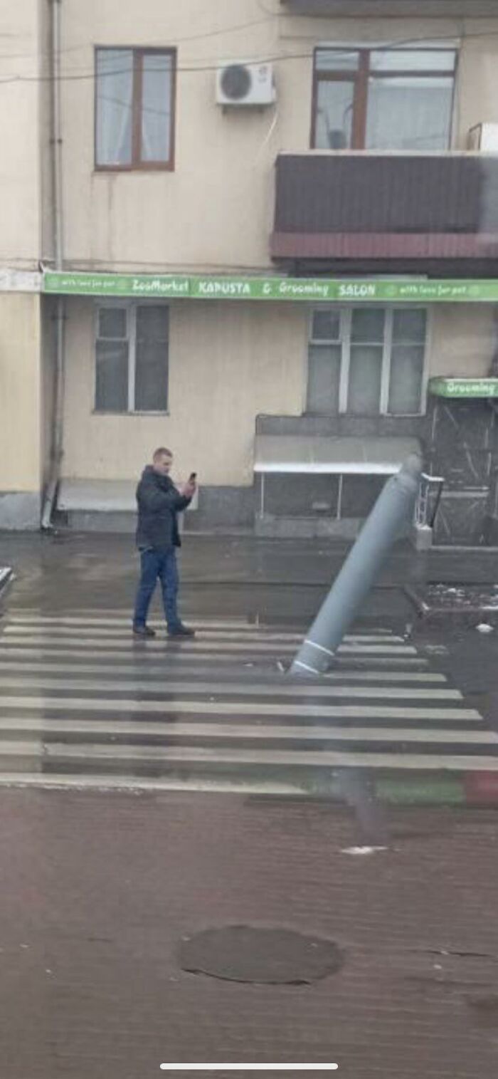 Hombre tomando foto a poste inclinado en paso peatonal, ilustrando razones por las que mujeres viven más que hombres.