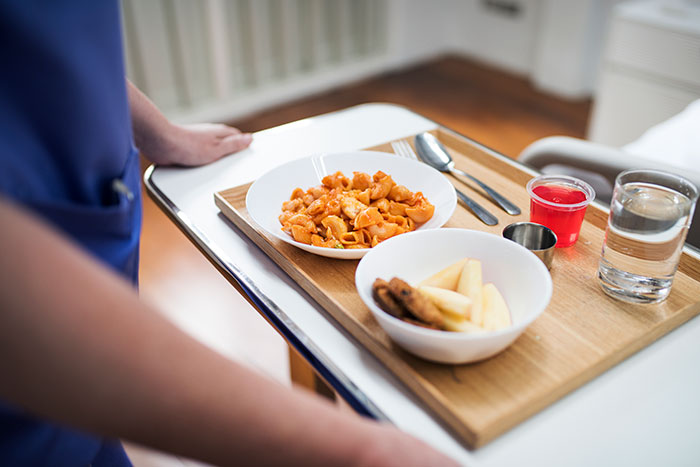 Trabajador sanitario llevando bandeja con comida, bebida y postre en habitación de paciente hospitalizado.