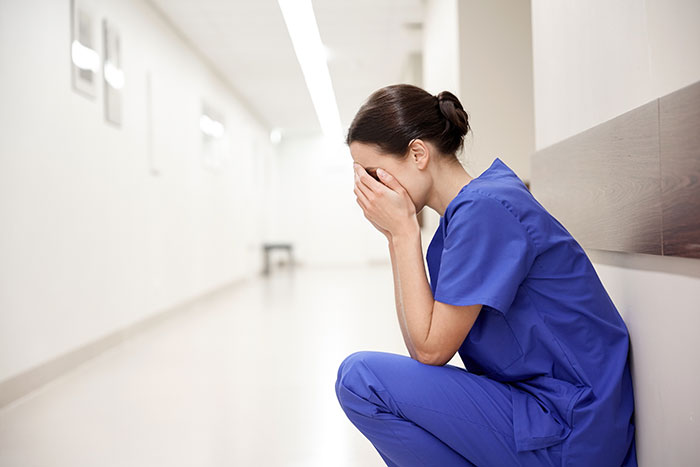 Enfermera en uniforme azul sentada en pasillo de hospital cubriéndose el rostro, momento difícil para trabajadores sanitarios.