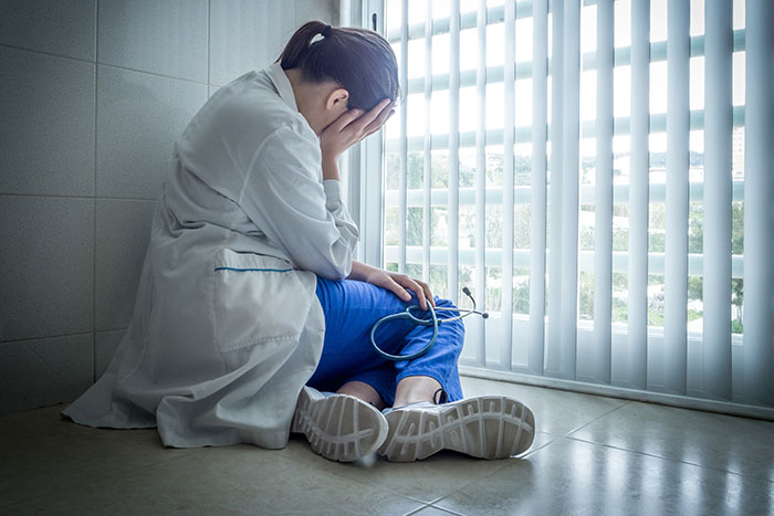 Trabajadora sanitaria sentada en el suelo junto a una ventana, mostrando cansancio y estrés en su jornada laboral.