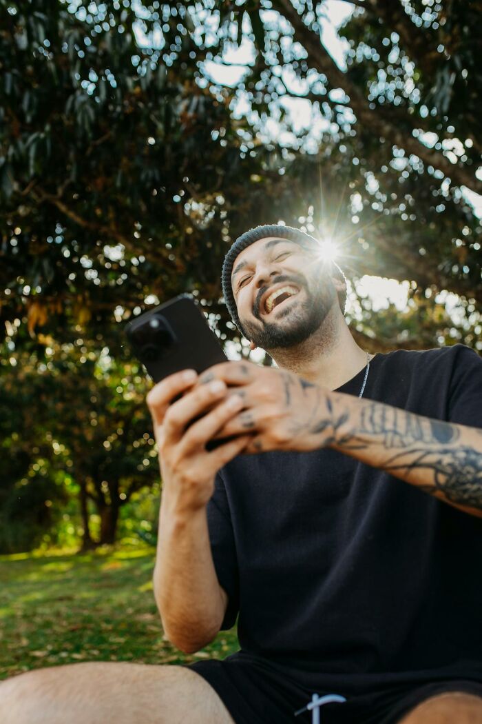 Hombre sonriente con tattoos usando celular al aire libre, representando momentos de corregir a la persona equivocada.