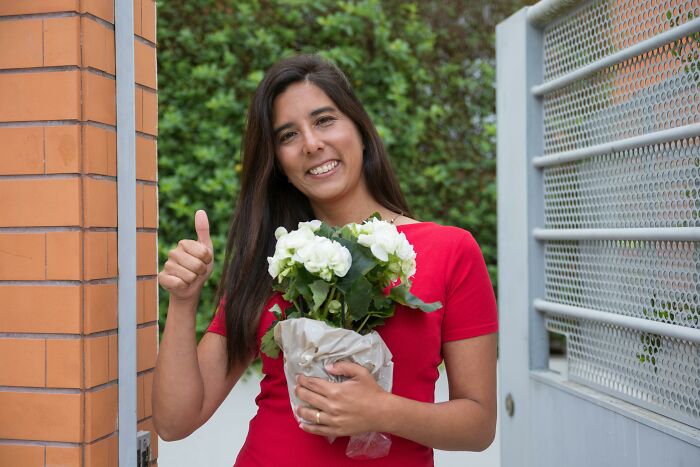 Mujer joven viviendo sola, sonriente con flores en mano y gesto de aprobación en la entrada de su hogar seguro.