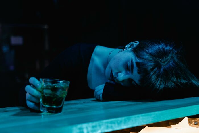 Mujer con expresión triste y cansada en un bar, sosteniendo un vaso, ambiente oscuro que sugiere conversaciones privadas terroríficas.